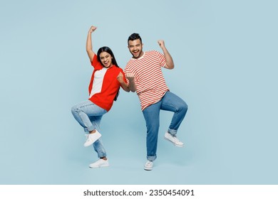 Full body young couple two friends family Indian man woman wear red casual clothes t-shirts together doing winner gesture celebrate clenching fists say yes isolated on plain blue cyan color background
