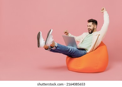 Full body young eacited overjoyed cool happy man in trendy jacket shirt sit in bag chair hold use work laptop pc computer do winner gesture isolated plain pastel light pink background studio 