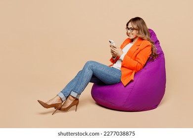 Full body young employee business woman corporate lawyer 30s wears classic formal orange suit glasses work in office sit in bag chair use mobile cell phone isolated on plain beige background studio - Powered by Shutterstock