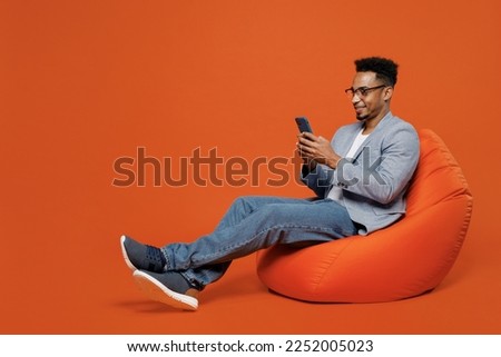 Full body young employee business man corporate lawyer wear classic formal grey suit shirt glasses work in office sit in bag chair hold use mobile cell phone isolated on plain red orange background