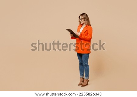 Full body young employee business woman corporate lawyer wear classic formal orange suit glasses work in office hold clipboard with paper account documents isolated on plain beige background studio