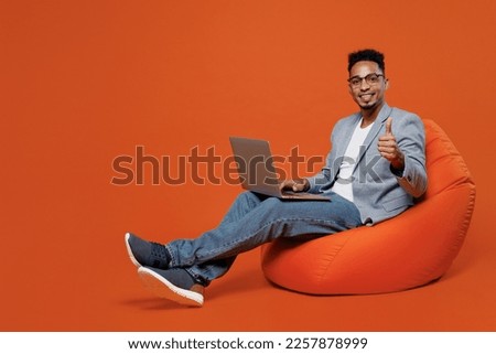 Full body young employee business man corporate lawyer wears formal grey suit shirt office sit in bag chair hold use work on laptop pc computer show thumb up isolated on plain red orange background