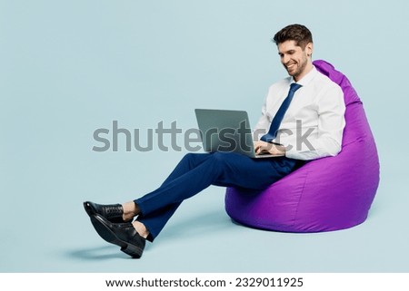 Full body young employee IT business man corporate lawyer wears classic formal shirt tie work in office sit in bag chair hold use work on laptop pc computer isolated on plain pastel blue background