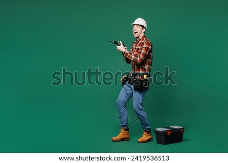 Full body young employee laborer man wear red shirt hardhat hat use hold electric drill isolated on plain green background. Instruments accessories for renovation apartment room. Repair home concept