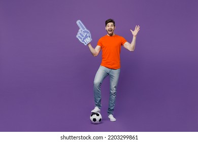 Full body young excited fun fan man he wear orange t-shirt cheer up support football sport team hold soccer ball fan foam glove finger up watch tv live stream isolated on plain dark purple background - Powered by Shutterstock