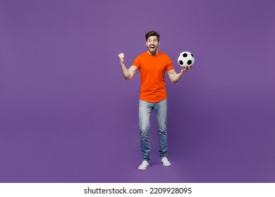 Full Body Young Excited Fun Fan Man He Wear Orange T-shirt Cheer Up Support Football Sport Team Hold In Hand Soccer Ball Watch Tv Live Stream Do Winner Gesture Isolated On Plain Dark Purple Background