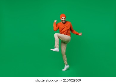 Full Body Young Excited Happy Man 20s Wear Orange Sweatshirt Hat Doing Winner Gesture Celebrate Clenching Fists Say Yes Raise Up Leg Isolated On Plain Green Background Studio People Lifestyle Concept.