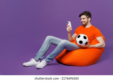 Full Body Young Fan Man He Wearing Orange T-shirt Cheer Up Support Football Sport Team Hold Soccer Ball Mobile Cell Phone Watch Tv Live Stream Sit In Bag Chair Isolated On Plain Dark Purple Background