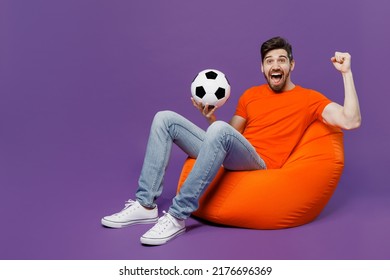 Full Body Young Fan Man He Wear Orange T-shirt Cheer Up Support Football Sport Team Hold Soccer Ball Watch Tv Live Stream Sit In Bag Chair Do Winner Gesture Isolated On Plain Dark Purple Background