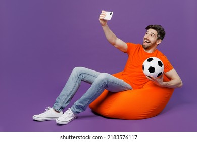 Full Body Young Fan Man He Wear T-shirt Cheer Up Support Football Team Hold Soccer Ball Watch Tv Live Stream Sit In Bag Chair Do Selfie Shot Mobile Cell Phone Isolated On Plain Dark Purple Background