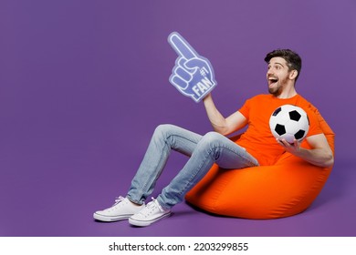 Full Body Young Fan Man Wear Orange T-shirt Cheer Up Support Football Sport Team Hold Soccer Ball 1 Fan Foam Glove Finger Up Watch Tv Live Stream Sit In Bag Chair Isolated On Plain Purple Background