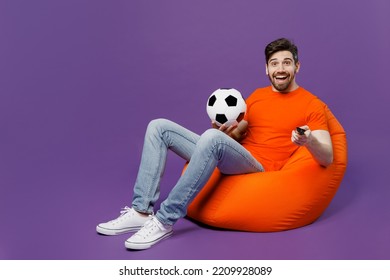 Full Body Young Fan Man He Wear Orange T-shirt Cheer Up Support Football Sport Team Hold Soccer Ball Remote Controller Watch Tv Live Stream Sit In Bag Chair Isolated On Plain Dark Purple Background