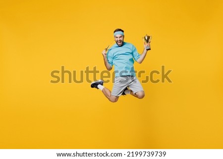 Full body young fitness trainer instructor sporty man sportsman in headband blue t-shirt golded medal hold winer cup jump high isolated on plain yellow background. Workout sport motivation concept.