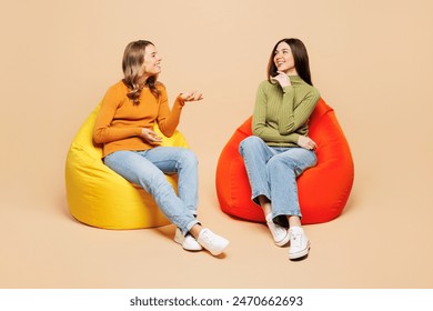 Full body young friends two women they wear orange green shirt casual clothes together sit in bag chair talk with each other isolated on plain pastel light beige background studio. Lifestyle concept