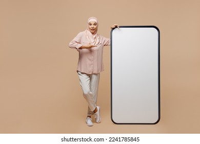 Full body young fun arabian asian muslim woman she in abaya hijab clothes point on big huge blank screen mobile cell phone isolated on plain light beige background People uae islam religious concept - Powered by Shutterstock