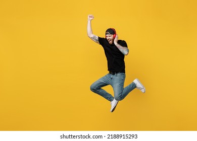 Full Body Young Fun Bearded Tattooed Man 20s He Wears Casual Black T-shirt Cap Headphones Listen To Music Jump High Do Winner Gesture Isolated On Plain Yellow Wall Background. People Lifestyle Concept