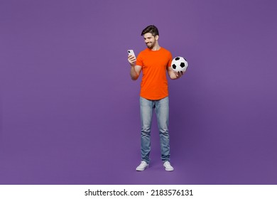 Full body young fun fan man 20s he wear orange t-shirt cheer up support football sport team hold in hand use mobile cell phone soccer ball watch tv live stream isolated on plain dark purple background - Shutterstock ID 2183576131