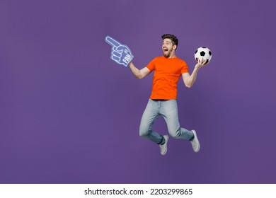 Full body young fun fan man wear orange t-shirt support football sport team hold in hand soccer ball 1 fan foam glove finger up watch tv live stream jump run isolated on plain dark purple background - Powered by Shutterstock