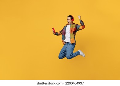 Full Body Young Fun Happy Cool Middle Eastern Man Wears Casual Shirt White T-shirt Headphones Listen Music Jump High Do Winner Gesture Hold Use Mobile Cell Phone Isolated On Plain Yellow Background