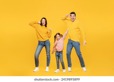 Full body young fun happy parents mom dad with child kid girl 7-8 years old wear pink sweater casual clothes do winner gesture dance show v-sign isolated on plain yellow background. Family day concept