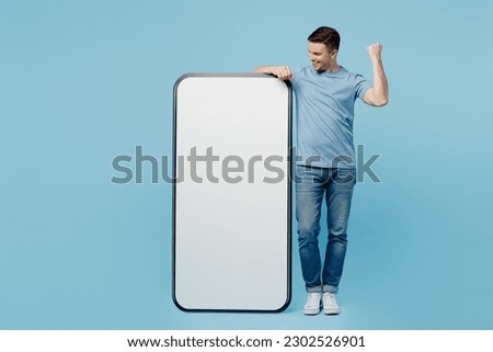 Full body young fun happy man wear casual t-shirt big huge blank screen mobile cell phone with workspace copy space mockup area do winner gesture isolated on plain pastel light blue cyan background