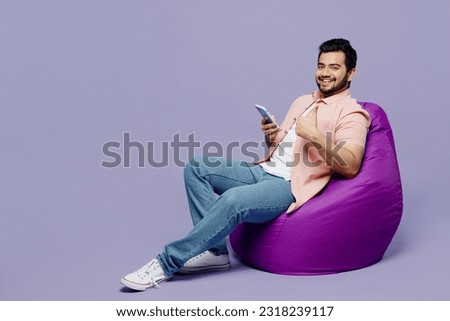 Full body young fun Indian man he wear pink shirt white t-shirt casual clothes use mobile cell phone show thumb up isolated on plain pastel light purple background studio portrait. Lifestyle concept