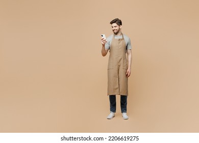 Full Body Young Fun Man Barista Barman Employee Wear Brown Apron Work In Coffee Shop Hold In Hand Use Mobile Cell Phone Isolated On Plain Pastel Light Beige Background. Small Business Startup Concept