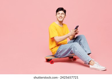 Full body young fun man he wears yellow t-shirt casual clothes sit on skateboard hold in hand use mobile cell phone isolated on plain pastel light pink background studio portrait. Lifestyle concept - Powered by Shutterstock