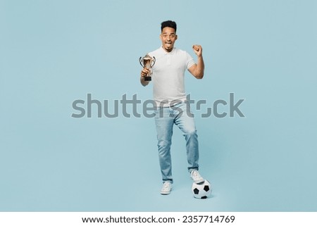 Full body young fun man fan wear t-shirt cheer up support football sport team hold in hand soccer ball golden cup watch tv live stream do winner gesture isolated on plain pastel blue color background