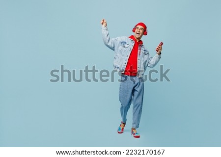 Full body young fun middle eastern man 20s he wear denim jacket red hat headphones listen music hold in hand use mobile cell phone sing song isolated on plain pastel lightblue cyan background studio