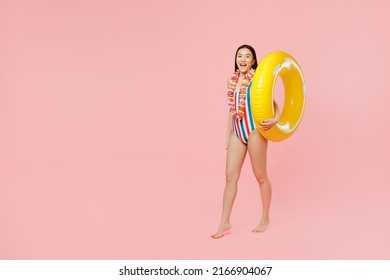 Full body young fun woman of Asian ethnicity in striped one-piece swimsuit hawaii lei hold inflatable ring walk go isolated on plain pastel pink background. Summer vacation sea rest sun tan concept