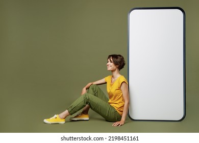 Full Body Young Fun Woman She Wear Yellow T-shirt Sit Near Big Huge Blank Screen Mobile Cell Phone With Workspace Copy Space Mockup Area Show Thumb Up Isolated On Plain Olive Green Khaki Background