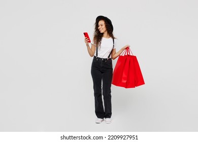 Full Body Young Fun Woman Wear T-shirt Hat Hold In Hand Red Paper Package Bags After Shopping Use Mobile Cell Phone Browsing Isolated On Plain Solid White Background. Black Friday Sale Buy Day Concept