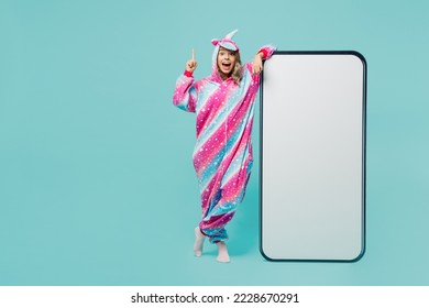 Full body young fun woman wear domestic costume with hoody and animals ears big huge blank screen mobile cell phone with area point index finger up isolated on plain pastel light blue cyan background - Powered by Shutterstock