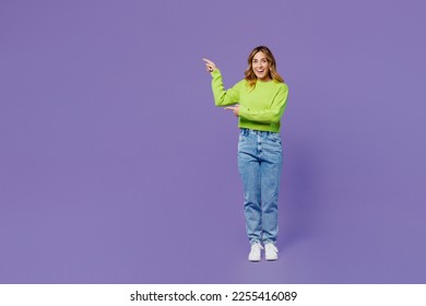 Full body young fun woman 30s she wearing casual green knitted sweater point index finger aside indicate on workspace area copy space mock up isolated on plain pastel purple background studio portrait