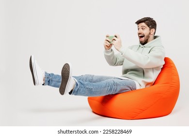 Full body young gambling man in mint hoody sit in bag chair use play racing app on mobile cell phone hold gadget smartphone for pc video games isolated on plain solid white background studio portrait