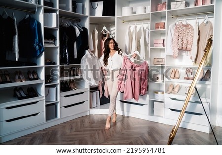 Full body of young happy barefoot female in trendy outfit standing near shelves in wardrobe and holding racks with garments