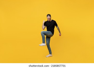 Full Body Young Happy Bearded Tattooed Man 20s He Wears Casual Black T-shirt Cap Doing Winner Gesture Celebrate Clenching Fists Say Yes Isolated On Plain Yellow Wall Background. Tattoo Translate Fun