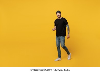 Full body young happy bearded tattooed man 20s he wears casual black t-shirt cap walking going strolling look camera isolated on plain yellow wall background studio portrait. People lifestyle concept