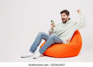 Full body young happy fun man wear mint hoody sit in bag chair hold in hand use mobile cell phone do winner gesture isolated on plain solid white background studio portrait. People lifestyle concept - Shutterstock ID 2253612457
