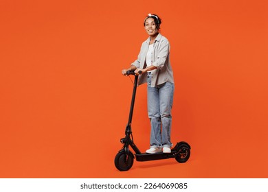 Joven cuerpo completo y feliz mujer de etnia afroamericana lleva una cámara de diseño de e-scooter con una banda de camisa gris aislada en un retrato de estudio de fondo naranja. Concepto de estilo de vida de la gente