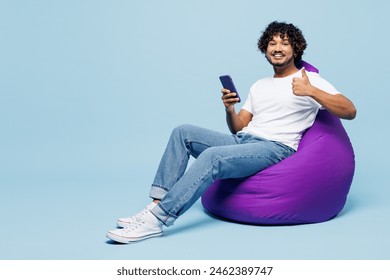 Full body young happy Indian man wear white t-shirt casual clothes sit in bag chair hold in hand use mobile cell phone show thumb up isolated on plain pastel light blue background. Lifestyle concept - Powered by Shutterstock