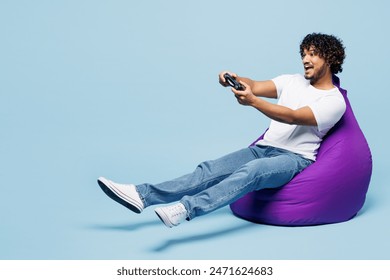 Full body young happy Indian man he wear white t-shirt casual clothes sit in bag chair hold in hand play pc game with joystick console isolated on plain pastel blue cyan background. Lifestyle concept - Powered by Shutterstock