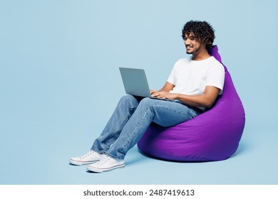 Full body young happy Indian man wear white t-shirt casual clothes sit in bag chair hold use work on laptop pc computer isolated on plain pastel light blue cyan background studio. Lifestyle concept - Powered by Shutterstock