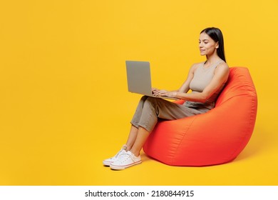 Full Body Young Happy Latin Woman 30s She Wear Basic Beige Tank Shirt Sit In Bag Chair Hold Use Work On Laptop Pc Computer Isolated On Plain Yellow Backround Studio Portrait. People Lifestyle Concept