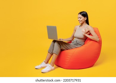 Full Body Young Happy Latin Woman 30s She Wearing Basic Beige Tank Shirt Sit In Bag Chair Hold Use Work On Laptop Pc Computer Waving Hand Talk On Video Call Isolated On Plain Yellow Backround Studio.