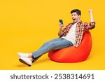 Full body young happy man wears red checkered shirt casual clothes sit in bag chair hold in hand use mobile cell phone do winner gesture isolated on plain yellow orange background. Lifestyle concept