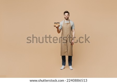 Full body young happy man barista barman employee wear brown apron work in shop hold craft paper brown cup coffee to go isolated on plain pastel light beige background. Small business startup concept