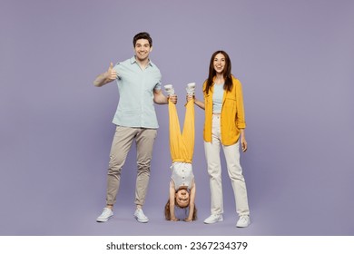 Full body young happy parents mom dad with kid daughter girl 6 years old wear blue yellow casual clothes holding child upside down show thumb up isolated on plain purple background. Family day concept - Shutterstock ID 2367234379