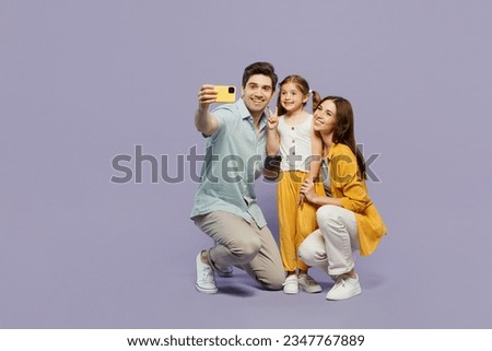 Full body young happy parents mom dad with child kid daughter girl 6 year old wear blue yellow casual clothes do selfie shot on mobile cell phone isolated on plain purple background Family day concept